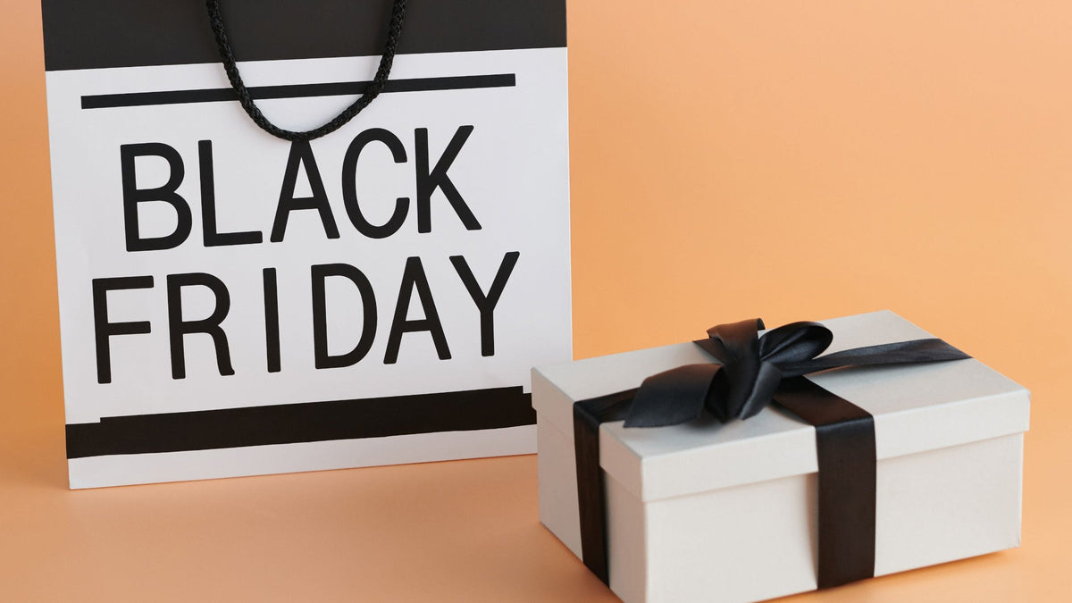 A black and white 'BLACK FRIDAY' shopping bag standing next to a white gift box tied with a black ribbon, set against a solid peach background.