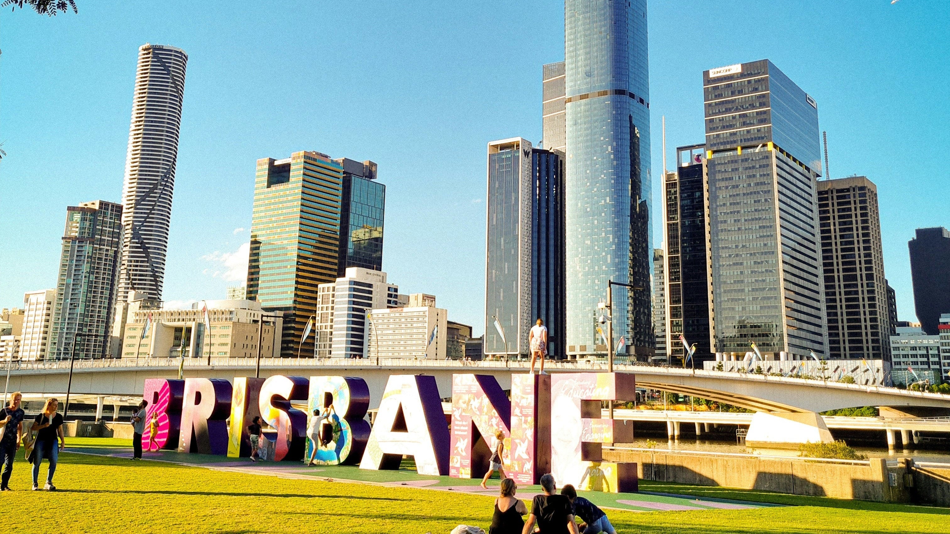A scenic outdoor photograph of the Brisbane skyline with the 'BRISBANE' letter sign in the foreground on a green lawn, under a clear blue sky.