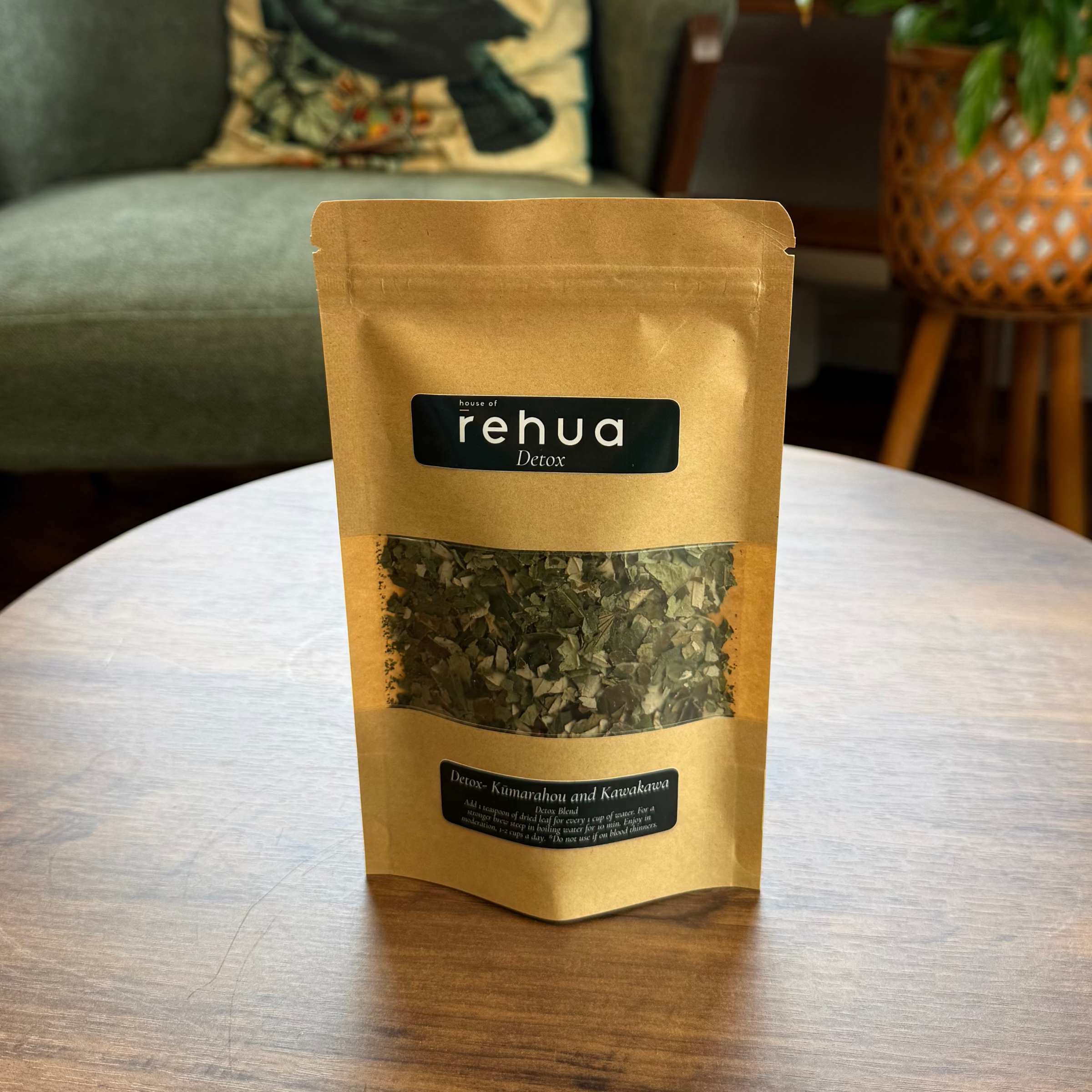 A stand-up pouch of rehua herbal detox tea sits on a round wooden table in a cozy, plant-filled room with a green armchair in the background.