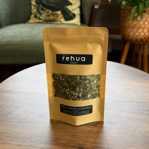 A stand-up pouch of rehua herbal detox tea sits on a round wooden table in a cozy, plant-filled room with a green armchair in the background.