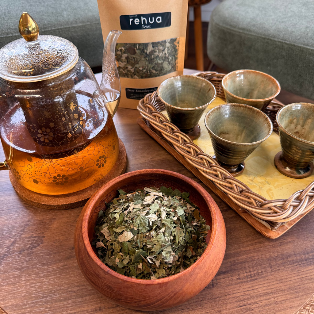 A House of rehua detox tea set arrangement on a wooden table featuring a glass teapot with amber tea, a bowl of loose detox tea leaves, a stand of four small ceramic cups on a tray, and a resealable pouch labeled 'rehua' in the background.