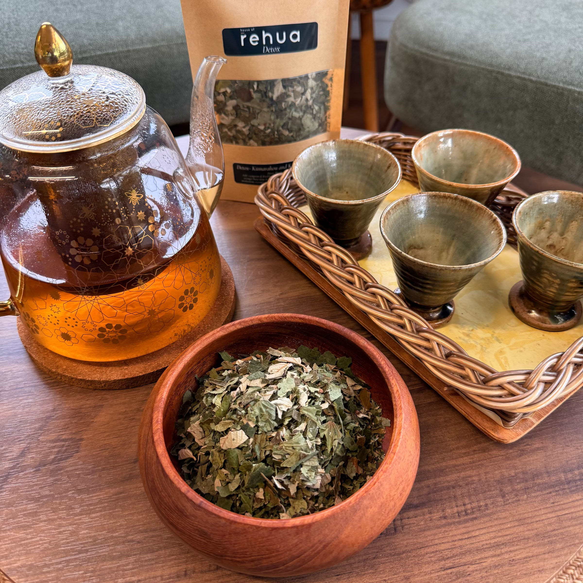A House of rehua detox tea set arrangement on a wooden table featuring a glass teapot with amber tea, a bowl of loose detox tea leaves, a stand of four small ceramic cups on a tray, and a resealable pouch labeled 'rehua' in the background.