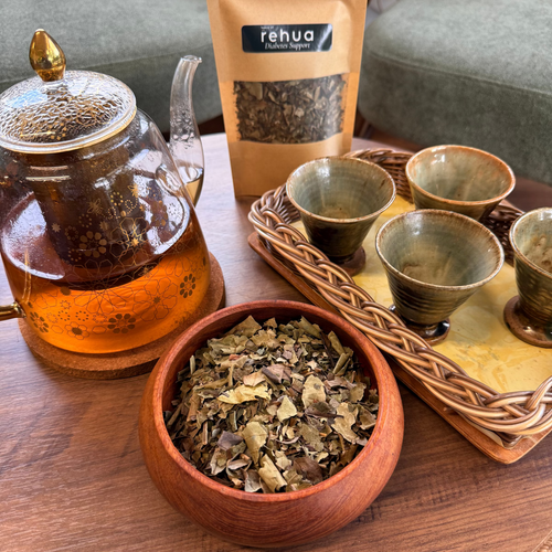 House of rehua diabetes support tea set arrangement on a wooden table featuring a glass teapot with amber tea, a bowl of loose Diabetes support  tea, and four small ceramic cups on a woven tray in a cozy living room setting.