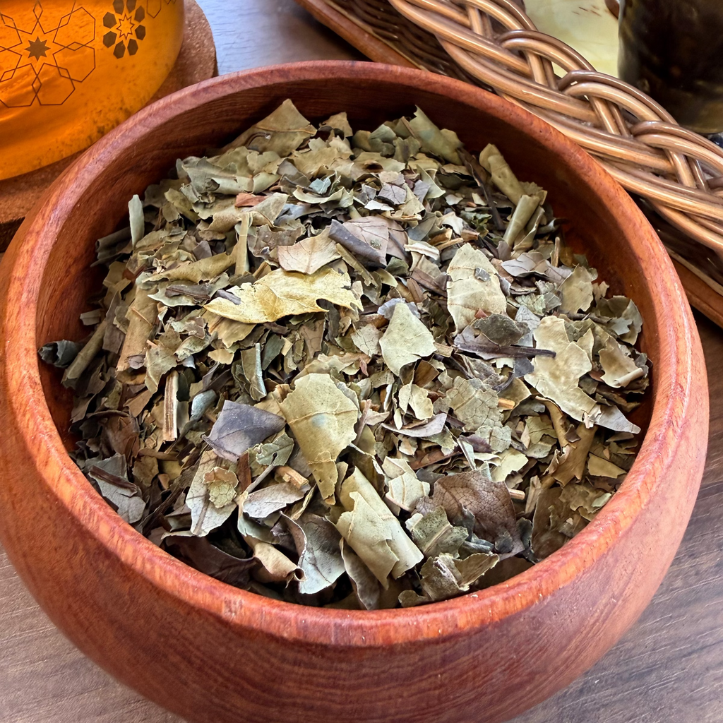 A close-up of loose Diabetes support  tea leaves in a round wooden bowl, set on a wooden table with woven trays and ceramic cups in the background.