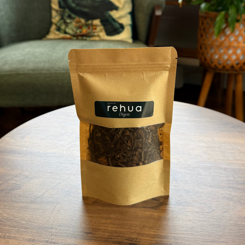 Pouch of house of rehua digest tea sits on a round wooden table in a cozy, plant-filled room with a green armchair in the background.
