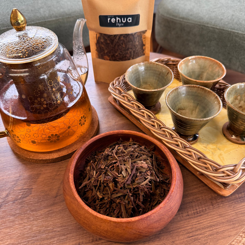 House of rehua Digest tea setup on a wooden table featuring a glass teapot filled with amber tea, a wooden bowl of loose Digest tea leaves, and four small ceramic cups on a woven tray, with a sofa in the background.