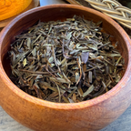 A wooden bowl filled with loose Digest tea leaves, set on a wooden table with a glass teapot and ceramic cups in the background.