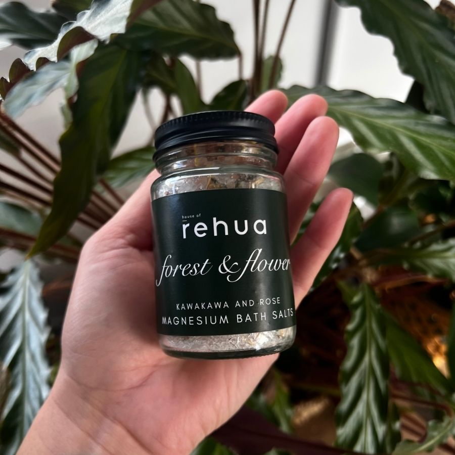 A hand holds a small glass jar of rehua 'forest & flower' magnesium bath salts, featuring a black label and black lid, set against a background of green houseplants.