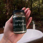 A hand holds a small glass jar of rehua Forest & Flower magnesium bath salts in front of a white bathtub with a forest background.