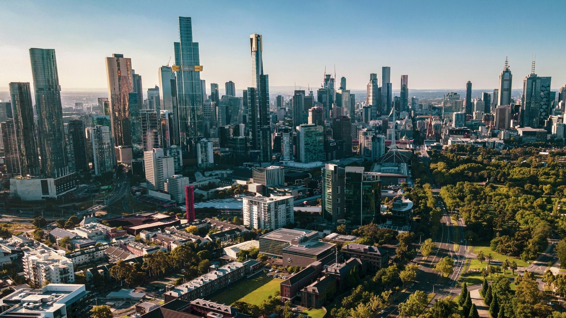 Aerial view of Melbourne city skyline with tall modern skyscrapers, low-rise buildings, green parks, and tree-lined streets under a clear blue sky.