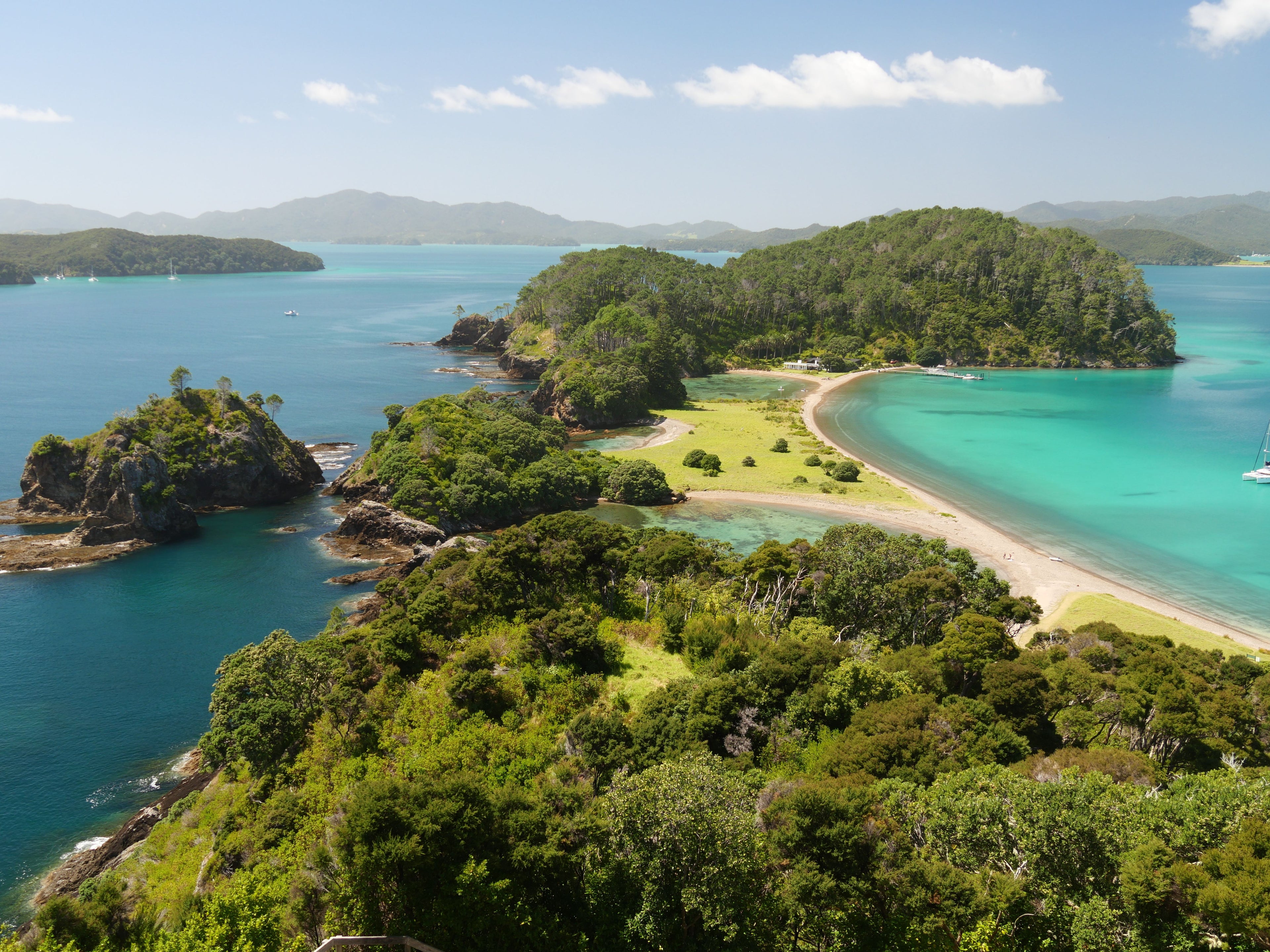Scenic aerial view of a tropical island coastline with turquoise and deep blue water, sandy beach, green forested hills, and distant mountains under a partly cloudy sky.