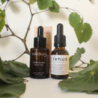 Two skincare oil bottles with droppers on a white surface surrounded by green leaves.