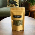 A stand-up pouch of House of Rehua Relax tea placed on a round wooden table in a cozy indoor setting with a green armchair, potted plants, and copper stills in the background.