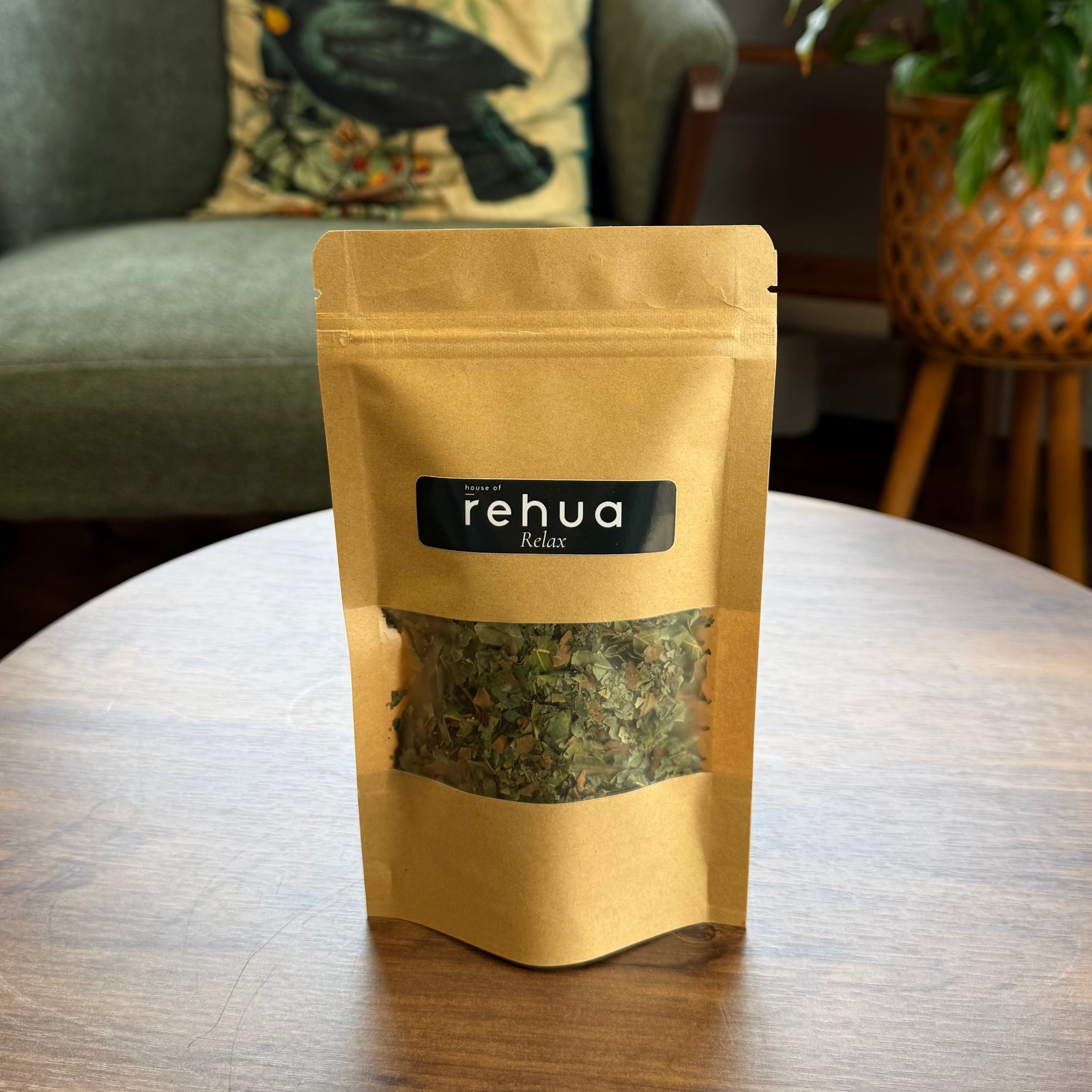 A stand-up pouch of House of Rehua Relax tea placed on a round wooden table in a cozy indoor setting with a green armchair, potted plants, and copper stills in the background.