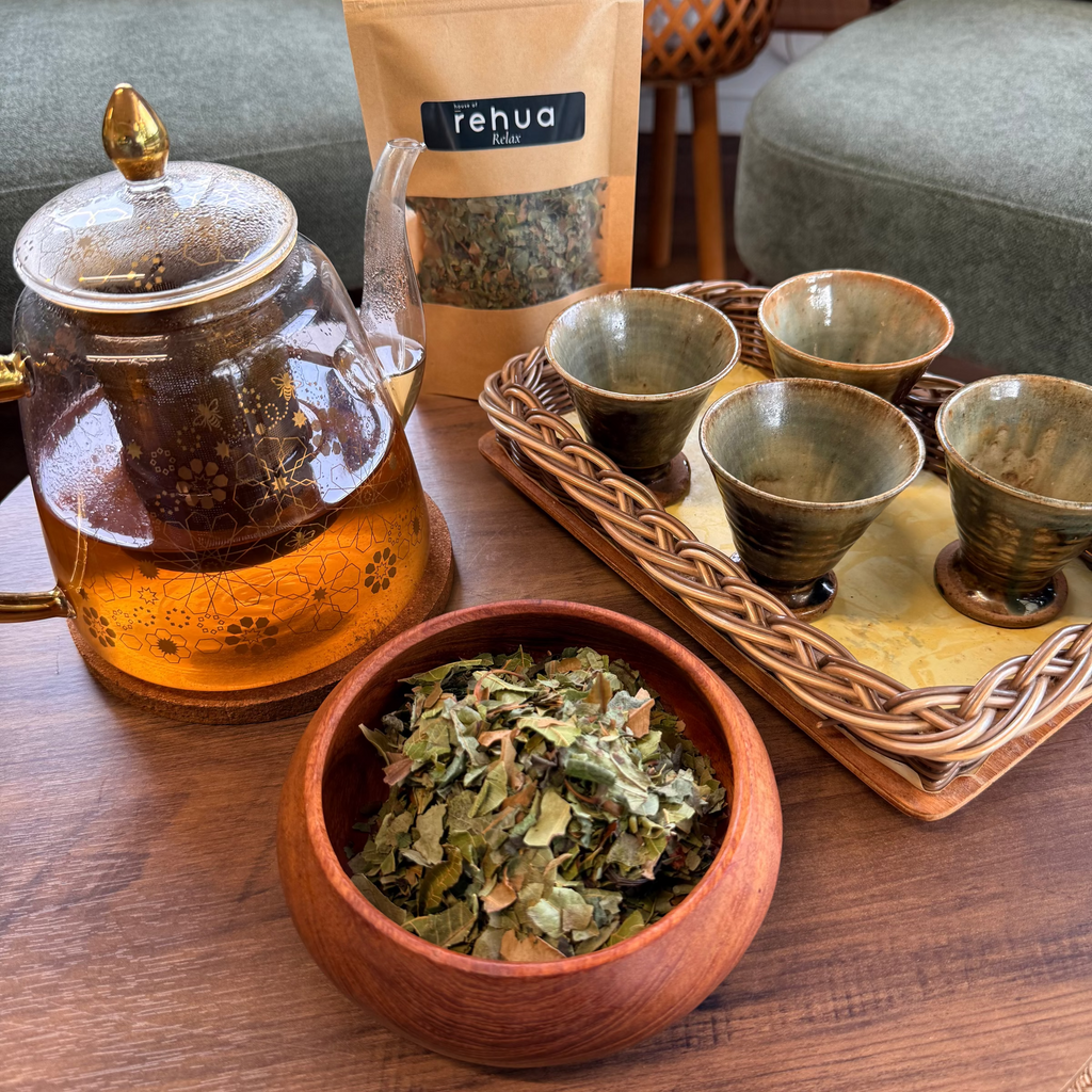 A tea preparation scene on a wooden table with a glass teapot filled with amber tea, a bowl of loose green tea leaves, a kraft pouch labeled 'Relax tea', and a tray of small ceramic cups in a home setting.