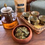 A tea preparation scene on a wooden table with a glass teapot filled with amber tea, a bowl of loose green tea leaves, a kraft pouch labeled 'Relax tea', and a tray of small ceramic cups in a home setting.