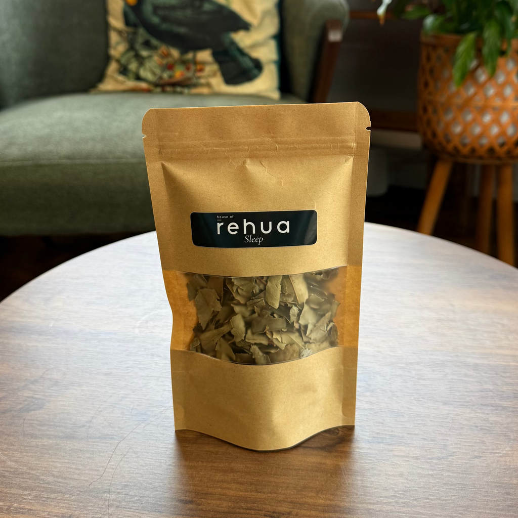 Pouch of house of rehua sleep tea, placed on a round wooden table in a cozy indoor setting with a green armchair, plants, and copper stills in the background.