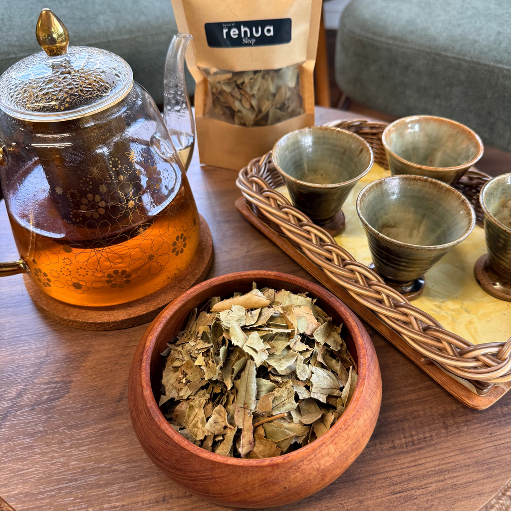 House of rehua Sleep tea setup on a wooden table featuring a glass teapot with amber liquid, a wooden bowl filled with Sleep tea leaves, a tray holding four small ceramic cups in a home interior.
