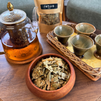 House of rehua Sleep tea setup on a wooden table featuring a glass teapot with amber liquid, a wooden bowl filled with Sleep tea leaves, a tray holding four small ceramic cups in a home interior.