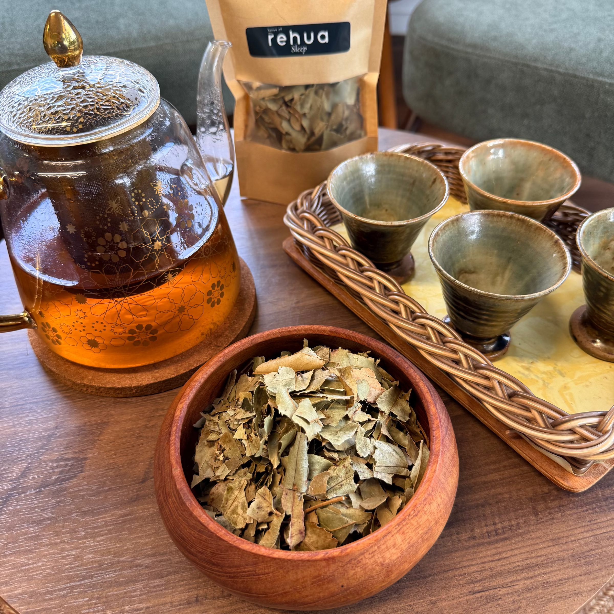 House of rehua Sleep tea setup on a wooden table featuring a glass teapot with amber liquid, a wooden bowl filled with Sleep tea leaves, a tray holding four small ceramic cups in a home interior.