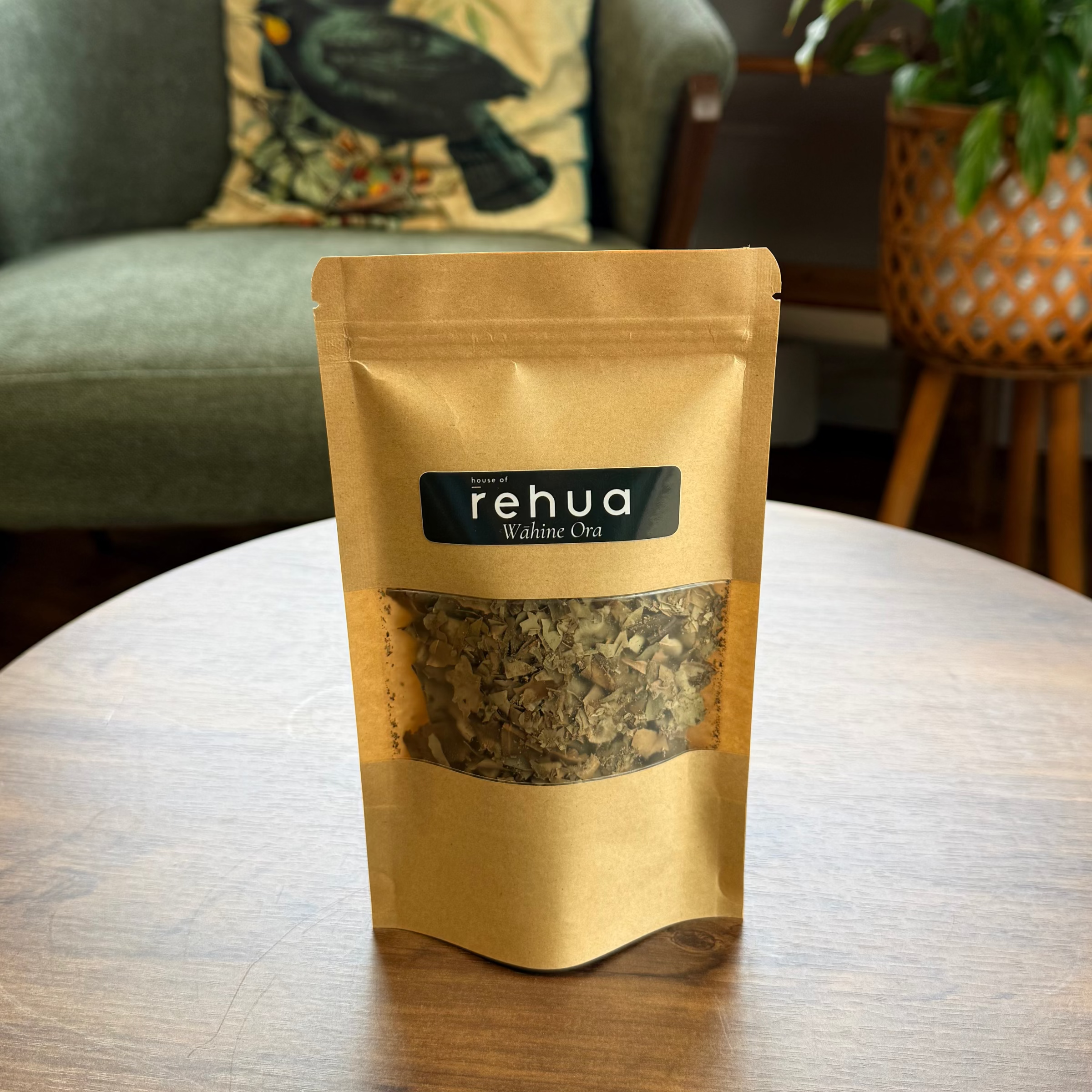 A stand-up pouch of House of Rehua Wahine ora tea sits on a round wooden table in a cozy, plant-filled room with a green armchair and decorative pillows in the background.
