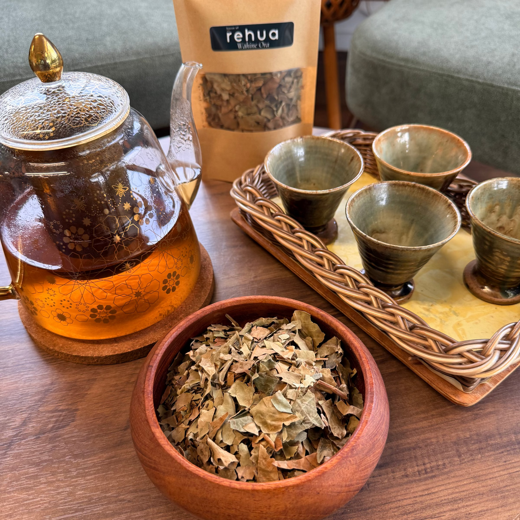 House of rehua Wahine ora tea setup on a wooden table featuring a glass teapot filled with amber tea, a wooden bowl of loose Wahine ora  tea leaves, a tray holding four small ceramic cups in a living room setting.