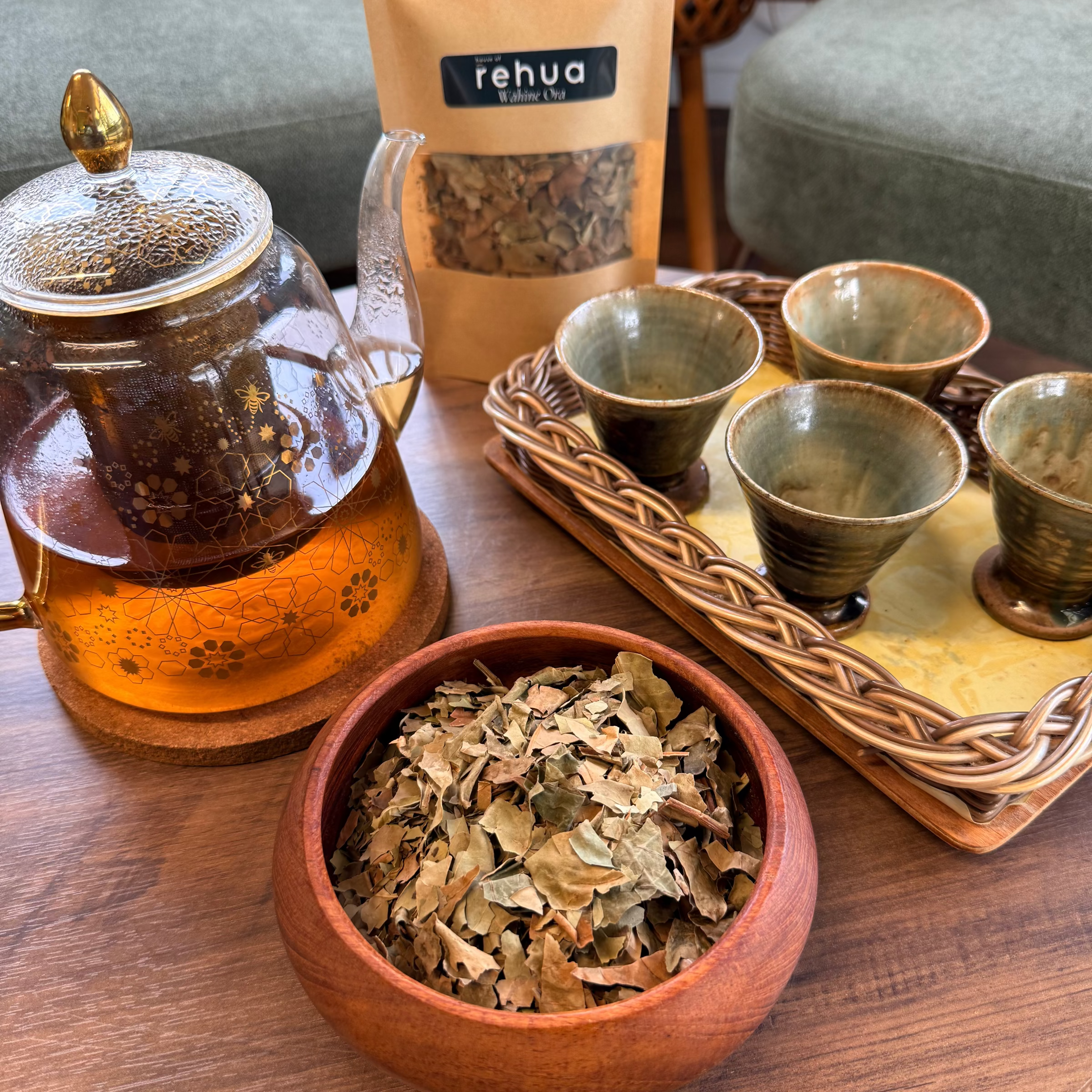 House of rehua Wahine ora tea setup on a wooden table featuring a glass teapot filled with amber tea, a wooden bowl of loose Wahine ora  tea leaves, a tray holding four small ceramic cups in a living room setting.