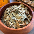 House of rehua Wahine ora tea leaves piled in a round wooden bowl on a tabletop, with a woven basket and amber glass vessel in the background.