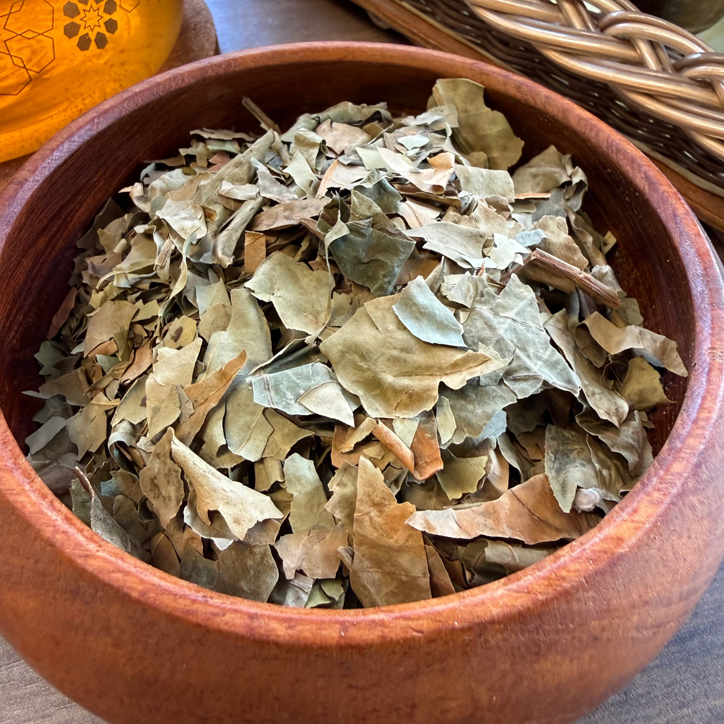 House of rehua Wahine ora tea leaves piled in a round wooden bowl on a tabletop, with a woven basket and amber glass vessel in the background.