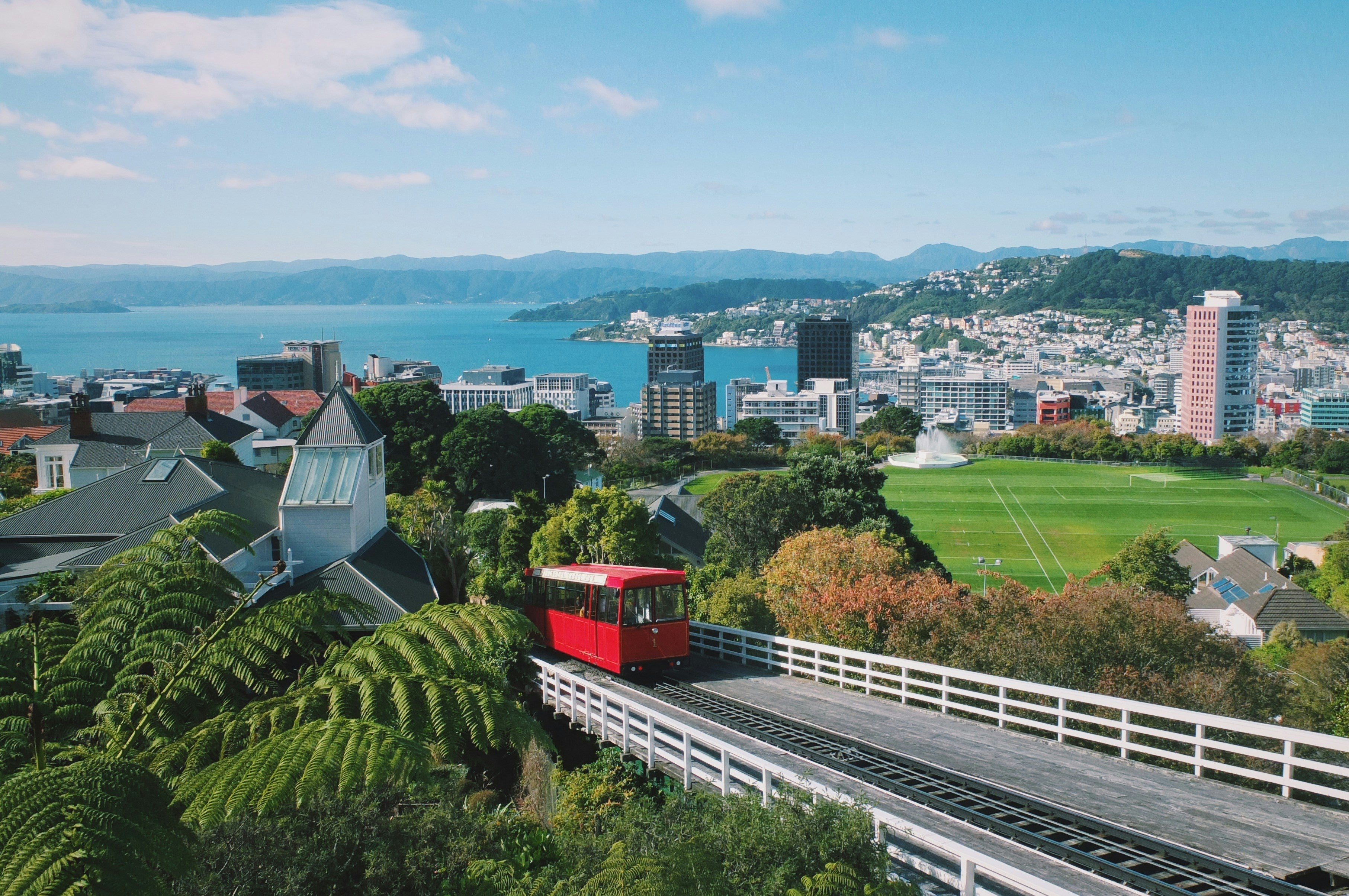 A scenic aerial view of a Wellington city with a red cable car traveling up a track through lush green trees, a large grassy field, and distant mountains and water under a blue sky.