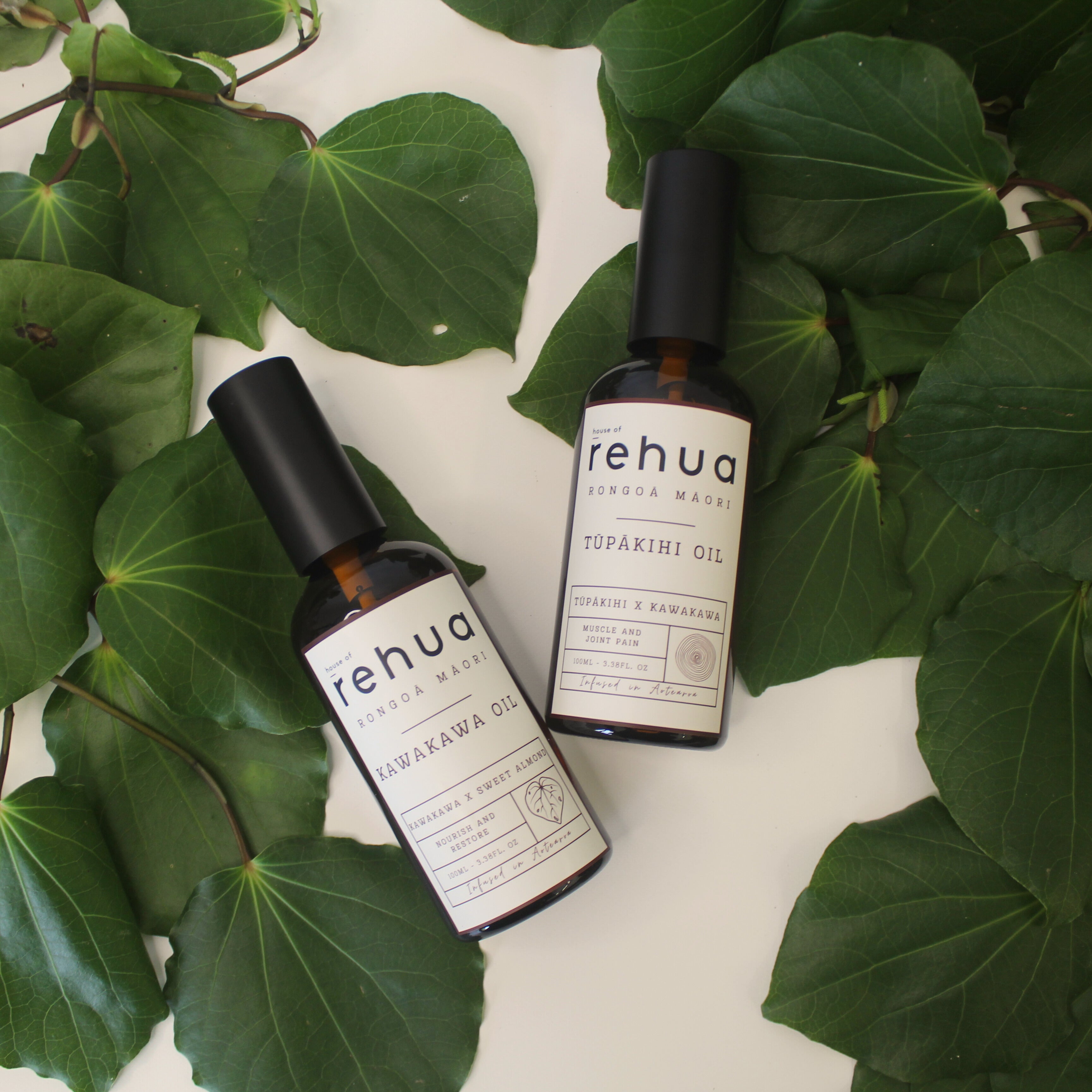 Two bottles of House Of Rehua Kawakawa and Tūpākihi Body Oil on a white surface with kawakawa leaves.