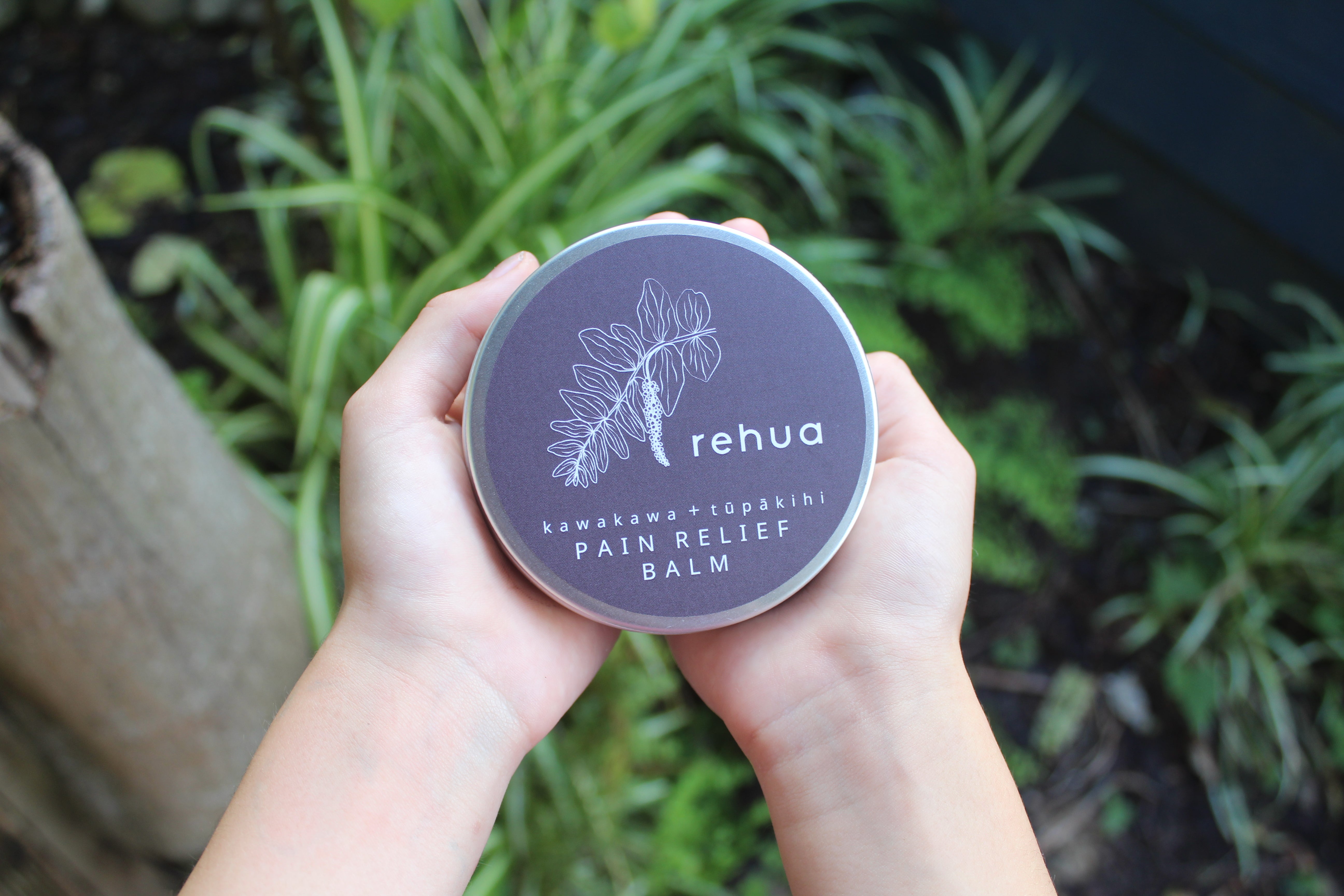 Hand holding a container of Rehua Pain Relief Balm with green plants in the background