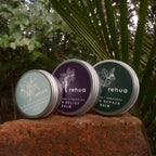 Three tins of House of Rehua balm on a stone surface with greenery in the background