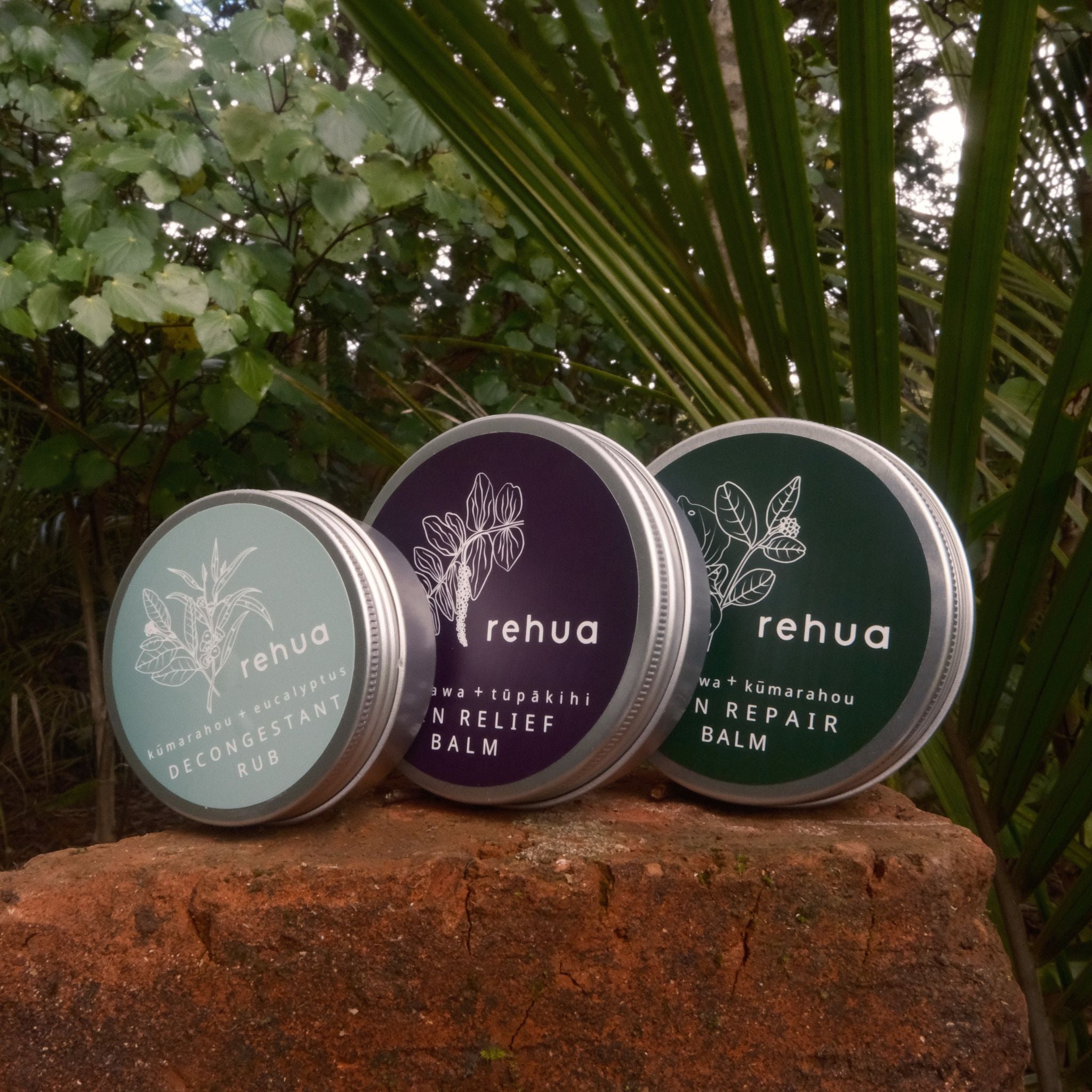 Three tins of House of Rehua balm on a stone surface with greenery in the background
