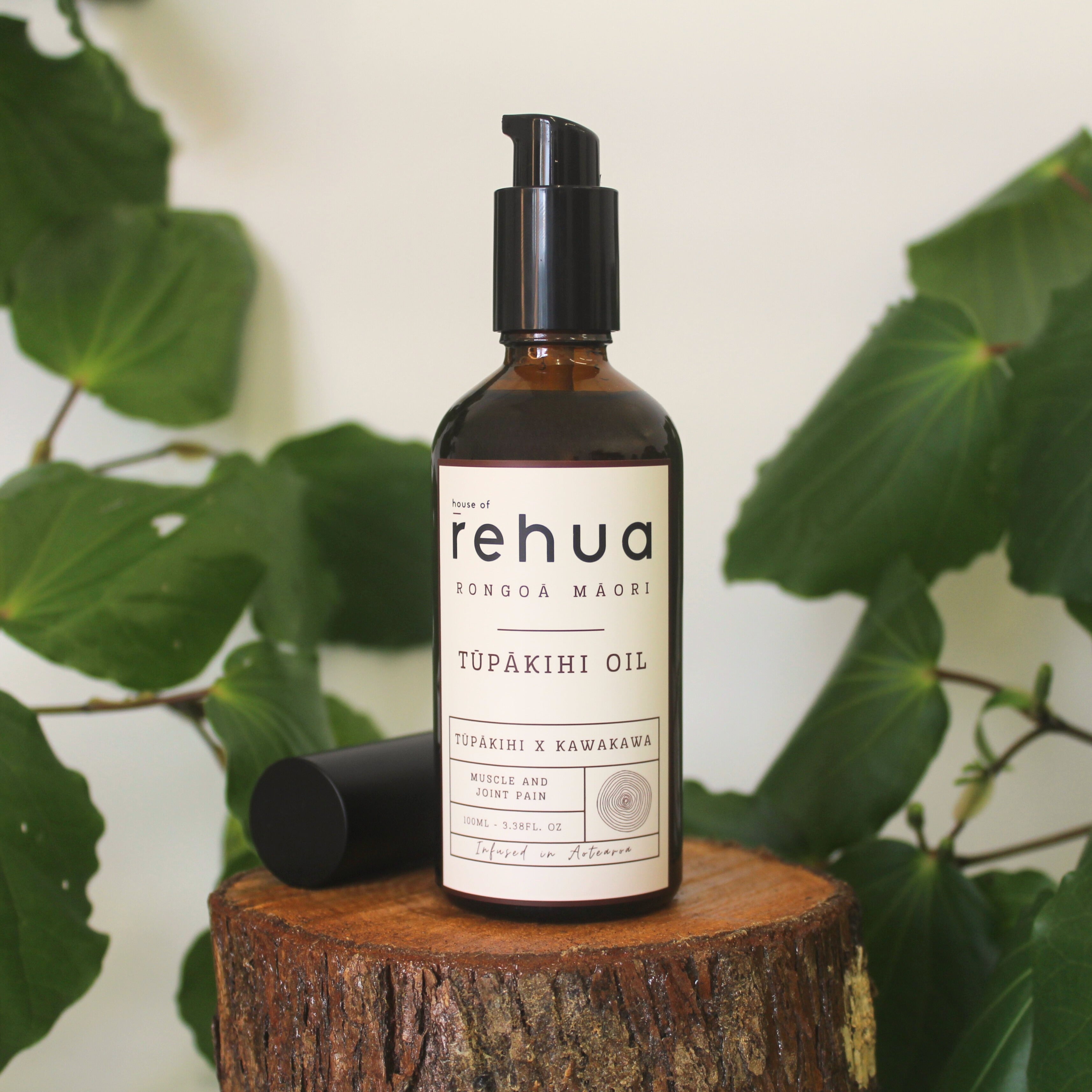 Bottle of House of Rehua Tūpākihi Body Oil on a wooden stump with kawakawa leaves in the background