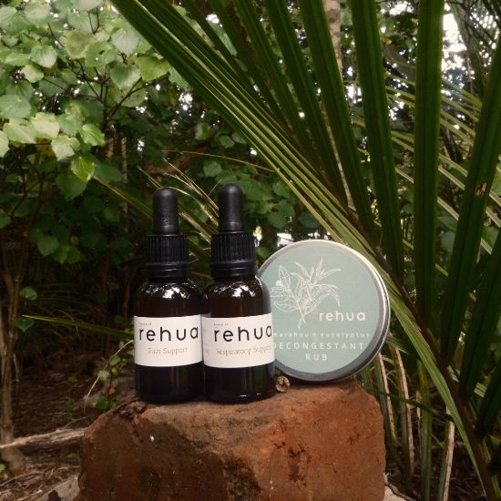 Two dropper bottles and a jar labeled 'rehua' on a stone surface with greenery in the background.