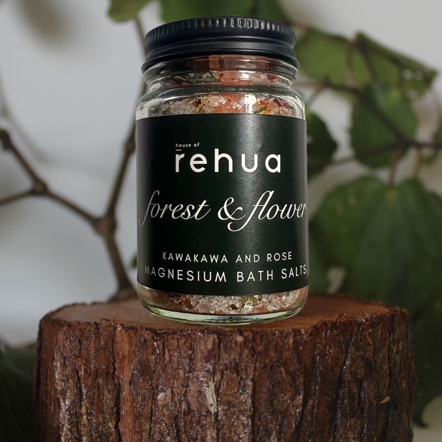 A glass jar of rehua 'forest & flower' magnesium bath salts with a black lid and black label, displayed on a rustic wooden log with soft-focus green leaves in the background.