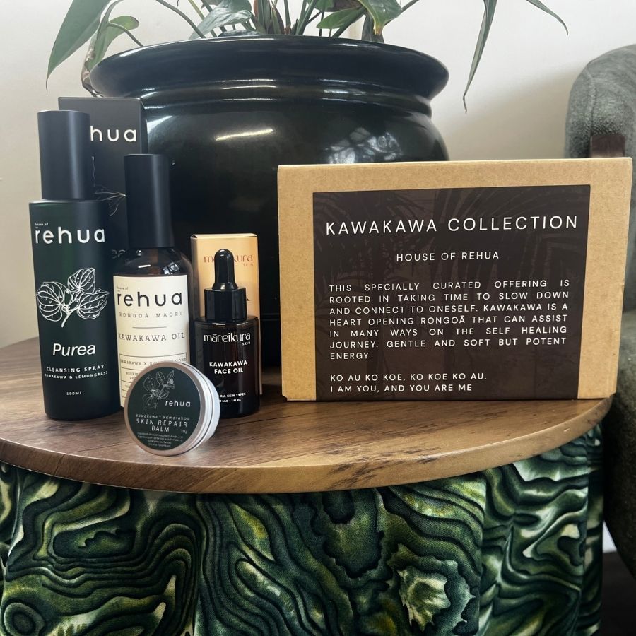 A curated skincare gift set from Rehua, including Purea Hydrating Spray, Kawakawa Face Oil, and a small jar, arranged on a wooden table next to a kraft paper box labeled 'KAWAKAWA COLLECTION HOUSE OF REHUA'.