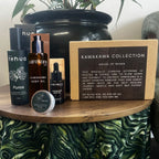 A curated skincare gift set from Kawakawa Collection House of Rehua displayed on a wooden table. The set includes a black Kawakawa Body Oil bottle, a black Kawakawa Face Oil dropper bottle, a black Rehua Purea spray bottle, a small round tin of Rehua Clay Mask, a kraft paper box labeled 'KAWAKAWA COLLECTION HOUSE OF REHUA', and a black Rehua box in the background, all arranged in front of a large black planter pot.
