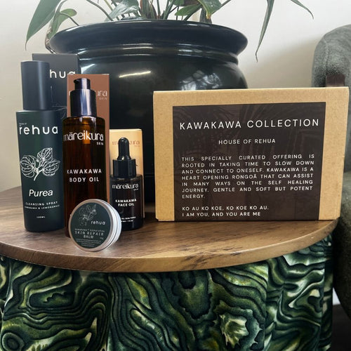 A curated skincare gift set from Kawakawa Collection House of Rehua displayed on a wooden table. The set includes a black Kawakawa Body Oil bottle, a black Kawakawa Face Oil dropper bottle, a black Rehua Purea spray bottle, a small round tin of Rehua Clay Mask, a kraft paper box labeled 'KAWAKAWA COLLECTION HOUSE OF REHUA', and a black Rehua box in the background, all arranged in front of a large black planter pot.