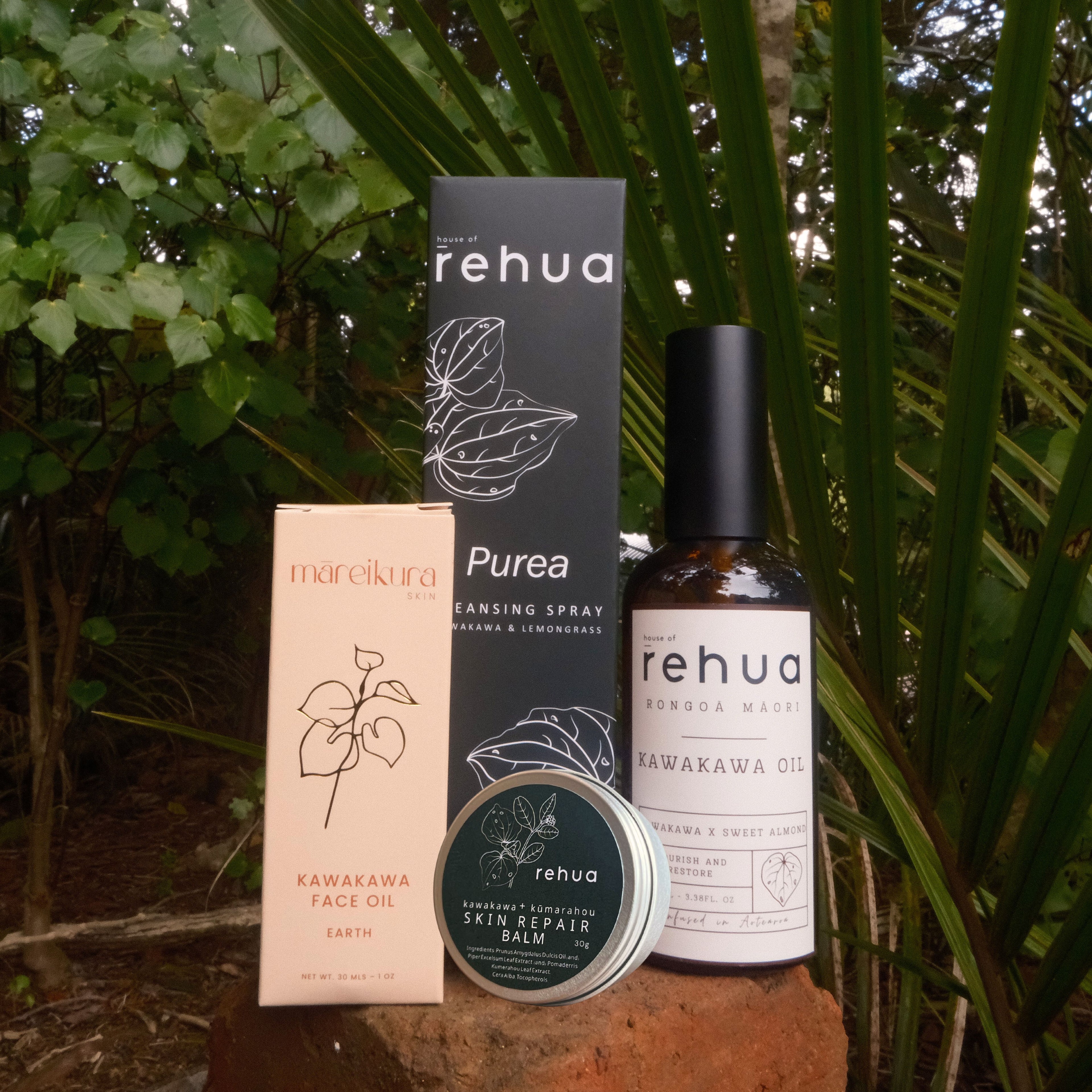 A skincare product set from rehua and mareikura arranged outdoors on a brick surface with palm leaves in the background. The set includes a dark charcoal box for Purea Cleansing Spray, a pale pink box for Kanakawaya Face Oil, a round tin of Skin Repair Balm, and a glass bottle of Rawakawaya Oil.