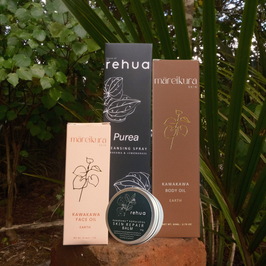 Three skincare products from rehua and mareikura are displayed outdoors on a brick surface with tropical green foliage in the background. The products include a black box of rehua Pureans Cleansing Spray, a brown box of mareikura Kawakaya Body Oil, a beige box of mareikura Kawakaya Face Oil, and a round tin of rehua Skin Repair Balm.