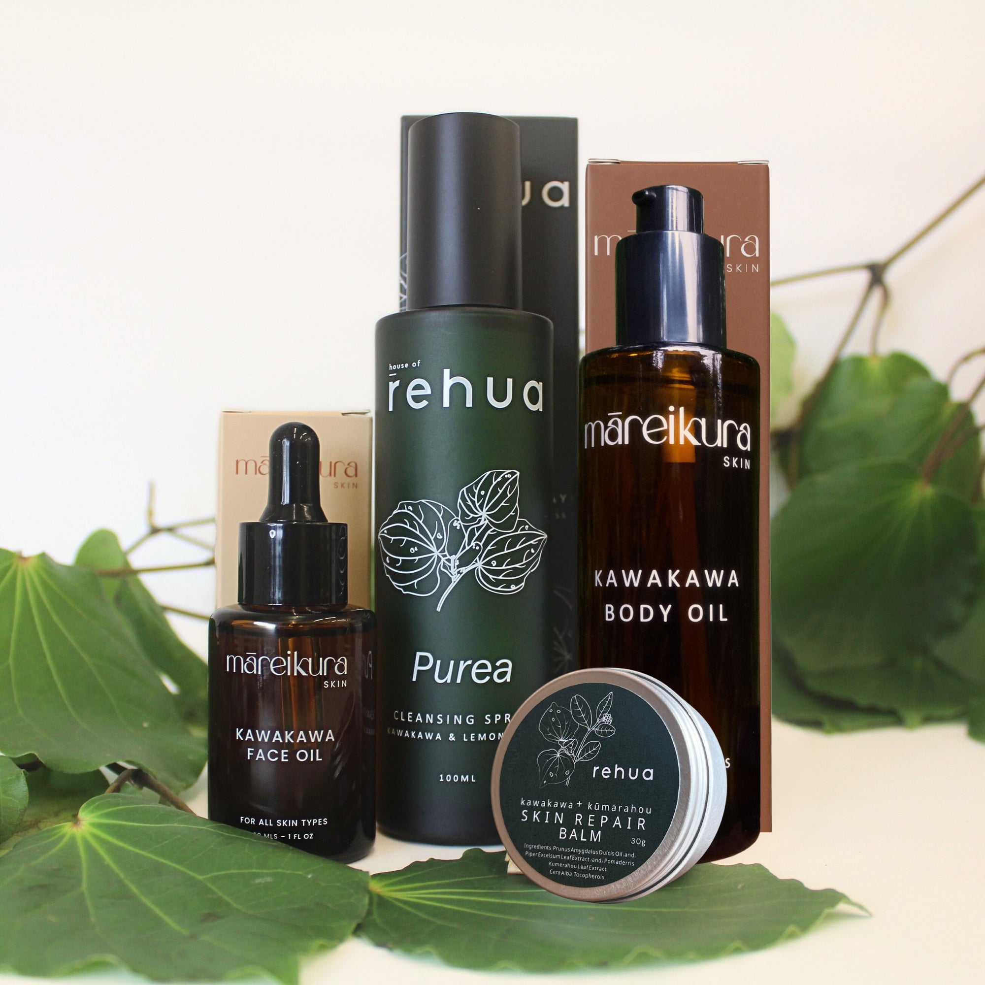 A skincare set from the brands rehua and mareikura skin, including face oil, cleansing serum, body oil, and skin repair balm, styled with green leaves on a light background.