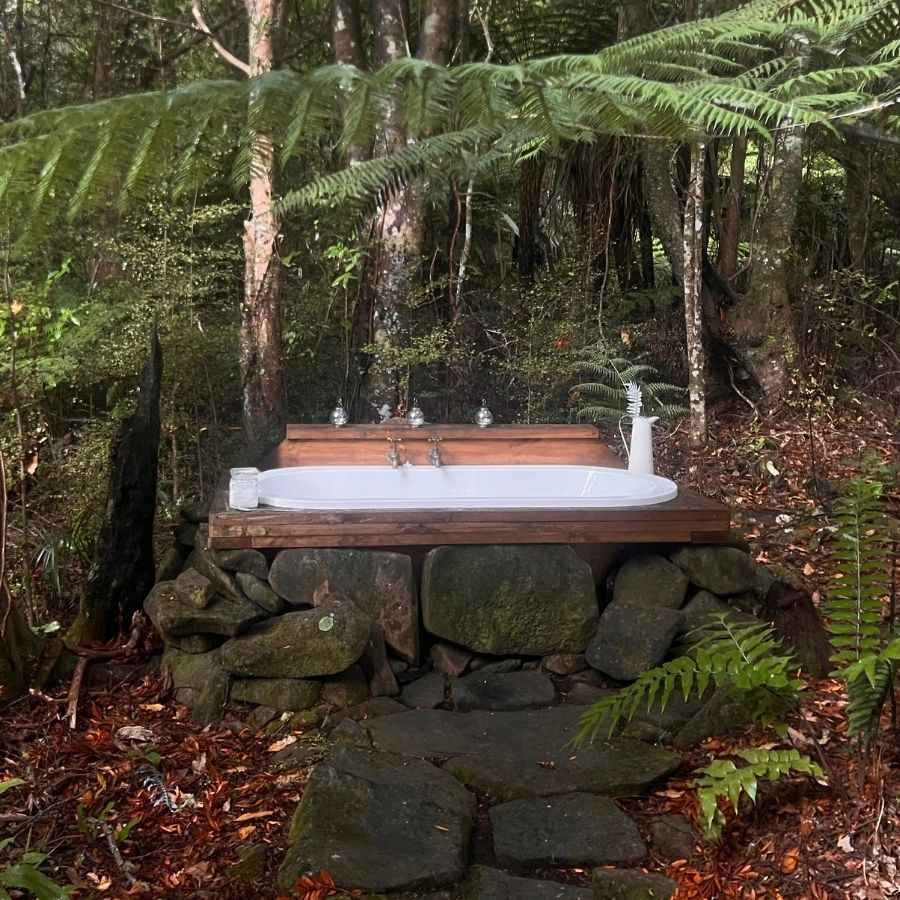 A white oval outdoor bathtub set on a wooden platform above a stone base, surrounded by a dense forest with ferns and fallen leaves.