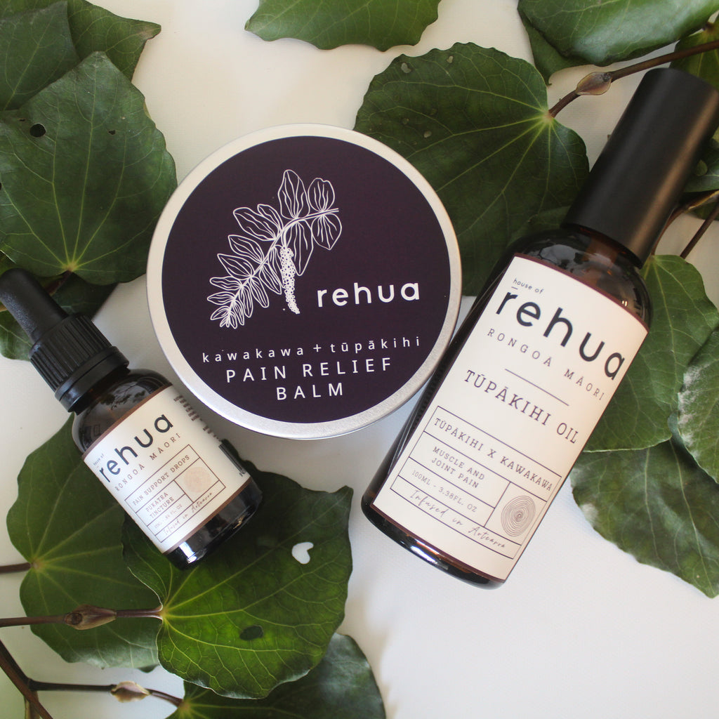 Rehua pain relief balm and Tupakihi oil bottles on a white surface with green leaves.