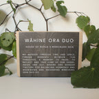Wooden sign with text 'W£ôhine Ora Duo' surrounded by green leaves on a light background